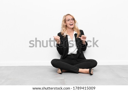 young blonde woman smiling cheerfully and casually pointing to copy space on the side, feeling happy and satisfied sitting on the floor