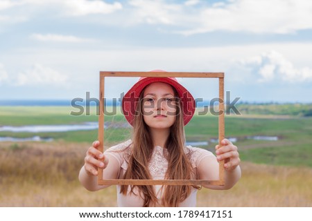 Girl in pink straw hat holds empty wooden picture frame in hands on nature landscape background. Portrait of a serious woman with long hair. Creative idea concept.