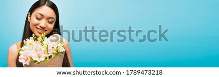 panoramic crop of happy asian girl smiling and holding flowers on blue