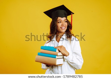 Graduate woman in a graduation hat on her head, with books stands on a yellow background. African American woman posing with smile. Graduation, university, college, distance education concept.