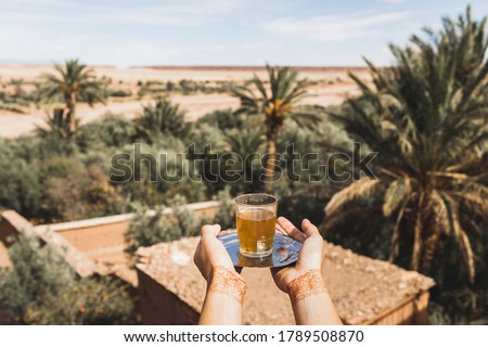 Woman hands with henna tattoo holding glass of traditional mint moroccan tea. Sand desert and palms on background.