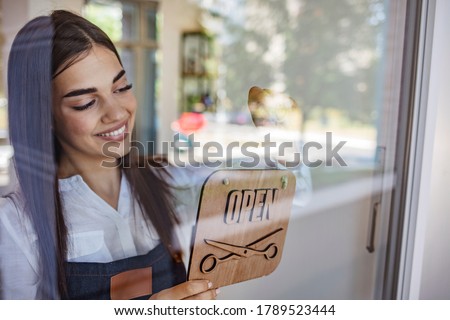 Portrait shot of a local business owner hanging an open sign on the retail glass window. Smiling female barber hanging open sign on door. Female owner is holding OPEN sign seen through glass.