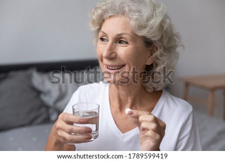Smiling happy healthy senior woman wake up sits on bed in bedroom alone holding glass of natural water taking daily pill for good health and senile disease prevention, memory meds, vitamins concept Royalty-Free Stock Photo #1789599419