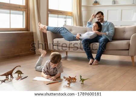 Smiling loving young bonding couple parents relaxing on comfortable sofa, watching little kid son drawing pictures in album, lying on wooden floor in living room, happy family weekend pastime.