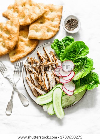 Mediterranean lunch - crispy italian focaccia, grilled turkey, green romaine salad and fresh vegetables on a light background, top view