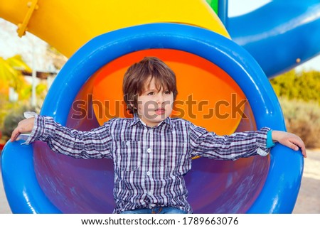 Handsome boy in plaid cowboy shirt goes down the round gutter. Beautiful sunny day. Children playground. Concept of photo advertising and happy childhood