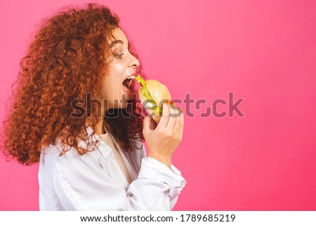 Junk food concept. Attractive Young curly Caucasian Woman Holding And Eating A Large Hamburger, Isolated over pink background. 