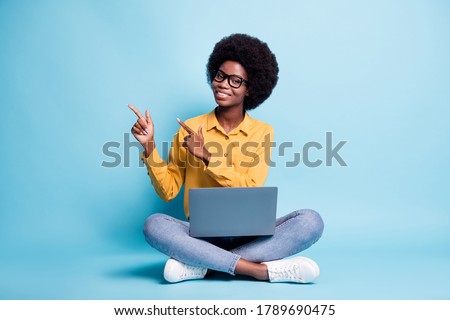 Full body size photo of black skin big volume hairstyle woman sit floor crossed legs hold netbook direct fingers empty space wear specs jeans yellow shirt isolated blue color background Royalty-Free Stock Photo #1789690475