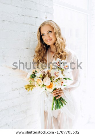 A blonde haired bride in a white peignoir posing in a light spare room with bouquet in her hands