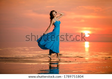 Young romantic girl walks relaxed by the water at sunset or sunrise. Beautiful woman in blue dress in boho style on background at sea. Sunrise over the ocean. Summertime scene