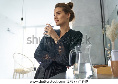 Low angle of smiling female in black dressing gown standing in kitchen with glass of water Royalty-Free Stock Photo #1789774949