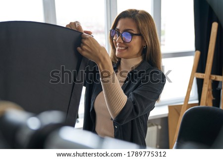 Cheerful female photographer in glasses is using modern lighting equipment for shooting in stylish office