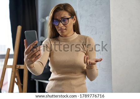 Beautiful female in glasses is holding smartphone and talking via camera online in art studio