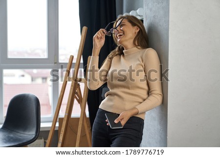 Jolly female with glasses is laughing in art studio while reading message on cell phone