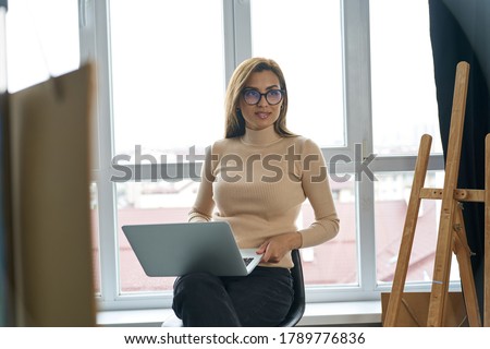 Trendy happy woman in glasses is sitting in fashion workshop and developing marketing on notebook
