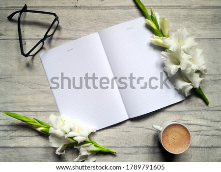 Home office desk workspace with gladioli flowers, coffee cup, open notebook and glasses. Blue wooden surface. Office day and working from home concept. Flat lay, from above, top view, copy space.