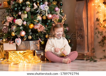 Merry Christmas and Happy Holidays! A cheerful girl with a rim of deer antlers sits near the New Year tree, on which balls and garlands hang, boxes with gifts stand.
Holiday concept.