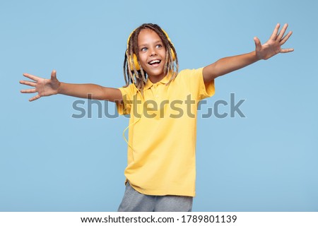 Joyful little boy with african dreads listening music in headphones dancing over blue background.
