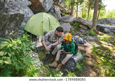 Father and his son sitting on stone and looking at pictures in mobile phone during their hiking