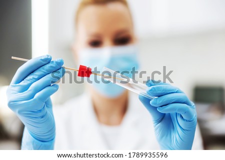 Blond female lab assistant in sterile uniform with rubber gloves and surgical mask on holding cotton swab. Laboratory interior. Royalty-Free Stock Photo #1789935596