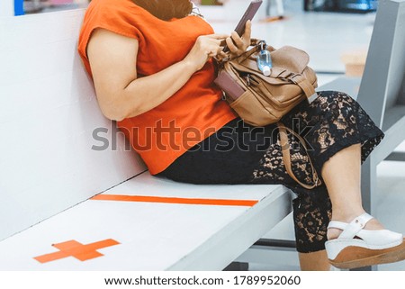Mini alcohol gel bottle to kill Corona Virus(Covid-19) hang on a leather shoulder bag of a woman wearing a mask while use smartphone with alternative seating mark for social distancing in the mall.