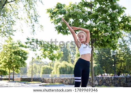 Young blond woman, wearing white top and black leggings,doing morning exercises in city park in summer. Training process outdoors. Three-quarter portrait of girl, stretching. Healthy lifestyle concept