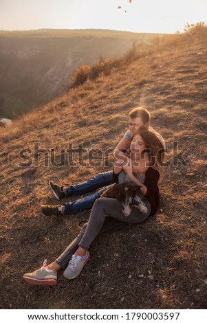 A young man hugs a beautiful girl from behind, the couple admire the sunset from the top of the hill. The lovers are sitting, hugging the little schnauzer. Happy family travels with a dog on an autumn
