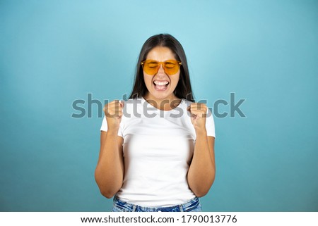 Young pretty woman wearing sunglasses over isolated blue background very happy and excited making winner gesture with raised arms, smiling and screaming for success. 