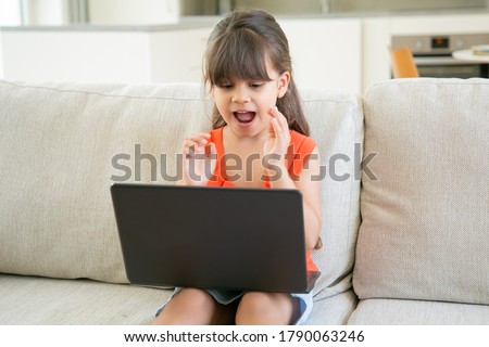Excited black haired little girl using laptop by herself. Cute child sitting on sofa and watching cartoons on pc. Internet and childhood concept
