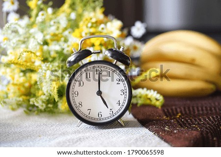 Blurred background of good summer day or morning with an alarm clock that shows 5 o'clock, a bouquet of wildflowers and bananas                            