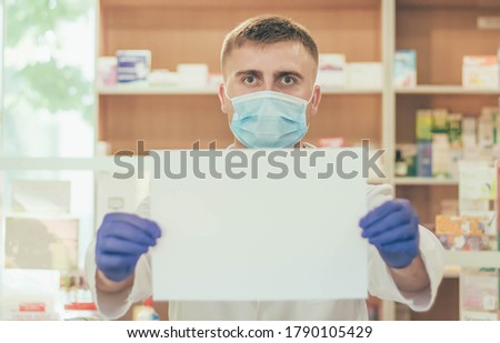 
A man pharmacist in a medical mask and a white coat against the background of a pharmacy counter holds a white piece of paper in front of him