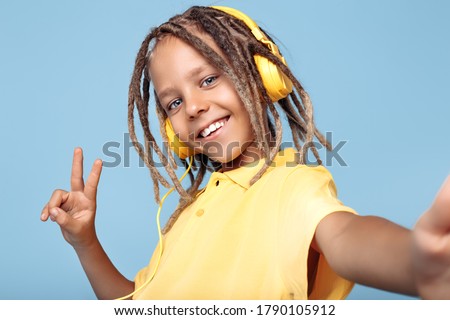 Positive little boy with african dreads wearing headphones taking selfie with smartphone over blue background. Peace.