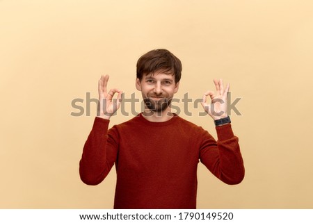 Photo of young man with beard wearing sweater, show okay signs, has smile, posing against yellow background.