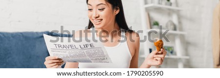 Panoramic crop of smiling asian woman holding piece of croissant while reading newspaper at home 