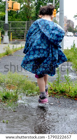 A young girl, pictured from behind in a blue rain poncho and pink shoes mid-stride on a rainy day, walks confidently through a puddle on the sidewalk, toward an intersection of a city street.