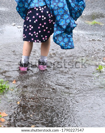 Closeup of the feet and legs of a young girl, pictured from behind, who walks through a deep puddle of rain water, wearing a blue rain poncho, pink shoes and a dress with a heart pattern.