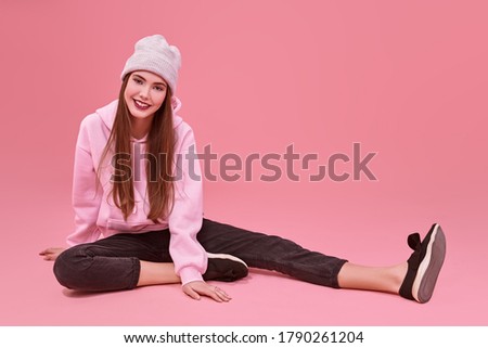 Youth fashion. Modern teen girl in a pink sweatshirt and a hat posing at studio on a pink background. Cosmetics and make-up. Full length portrait.