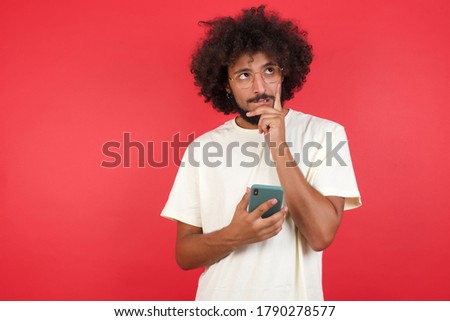 Image of a thinking dreaming young handsome man posing isolated over red wall using mobile phone and holding hand on face. Taking decisions and social media concept.