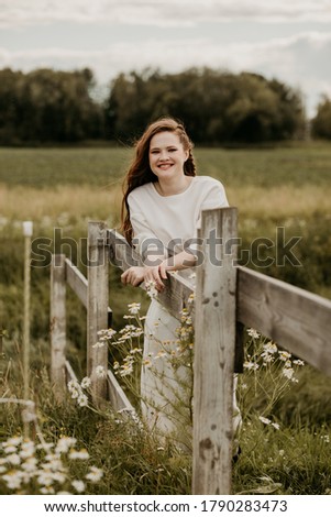 Beautiful young red-haired woman stands next to a wooden hedge in a field among daisies in summer. Image with selective focus