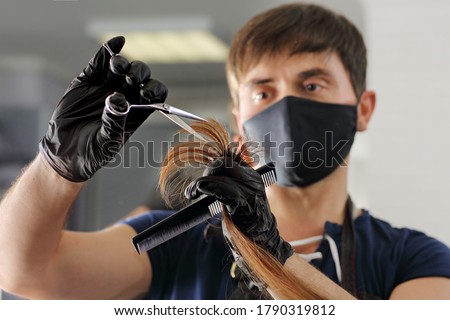 A hairdresser cuts the long hair of a young girl. A Barber in black nitrile gloves and a reusable mask uses scissors and a comb. Royalty-Free Stock Photo #1790319812