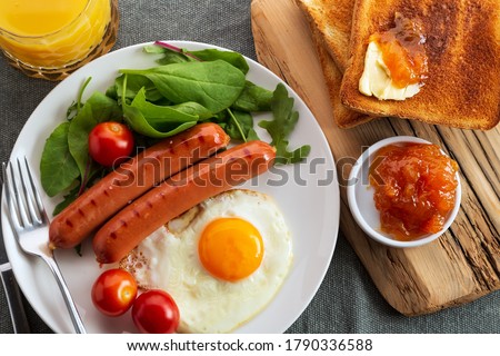 Table with food, breakfast or brunch concept. Top view. Fried egg, grilled sausages, vegetables, herbs, toast with jam and butter. Royalty-Free Stock Photo #1790336588