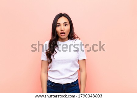 young latin pretty woman looking very shocked or surprised, staring with open mouth saying wow against flat wall