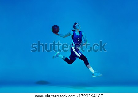 In high flight. Young caucasian female basketball player training, prcticing with ball isolated on blue background in neon light. Concept of sport, movement, energy and dynamic, healthy lifestyle.