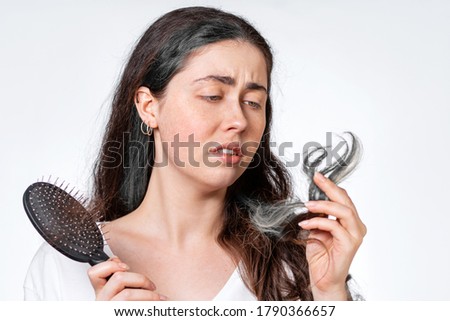 A saddened brunette woman holds a comb with her hair falling out and looks at the gray tips of her hair. White background. The concept of hair loss and gray hair Royalty-Free Stock Photo #1790366657