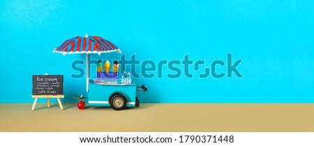 Ice cream toy cart with blue red umbrella. Assortment of ice cream menu black chalkboard. Summer vacation concept. Blue sandy beach background. copy space.