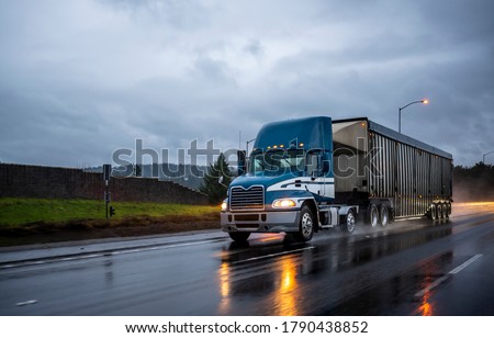 Big rig day cab bonnet blue semi truck with roof spoiler transporting commercial cargo in covered corrugated bulk semi trailer running on the evening wet glossy slippery road with raining weather Royalty-Free Stock Photo #1790438852