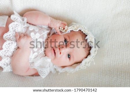 A little cute 2 weeks old newborn baby kid girl dressed in a lace top and cap is lying in the white background, touching her face