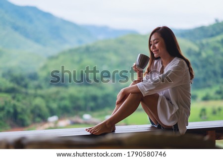 Portrait image of a beautiful asian woman holding and drinking hot coffee on balcony , looking at mountains and green nature 