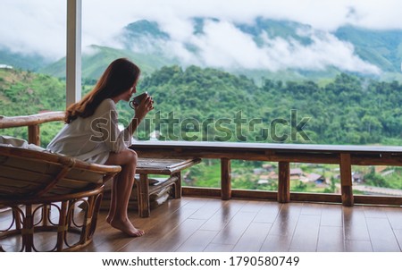 Portrait image of a beautiful asian woman holding and drinking hot coffee , sitting on balcony and looking at mountains and green nature 