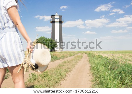 Girl in stripes overall with bunck of wheat and straw hat in right hand goes to black and white lighthouse y dirt road.